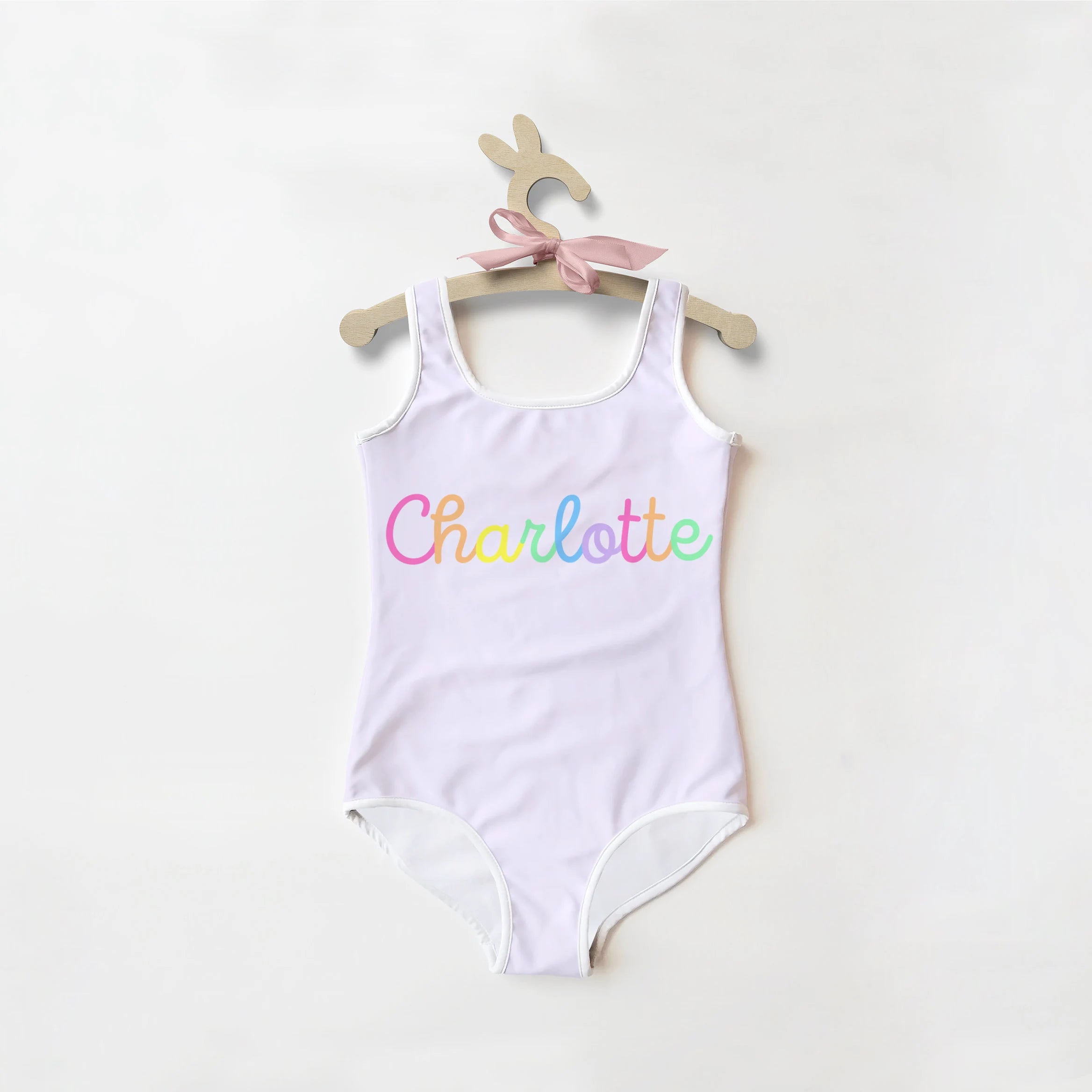 A white one-piece girls swimsuit with custom rainbow charlotte name hanging on a wooden bunny-shaped hanger