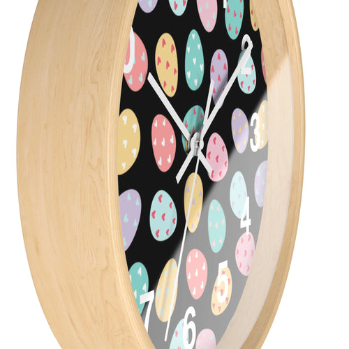 A round silent wood wall clock with a light wood frame and black face decorated with pastel easter eggs and hearts