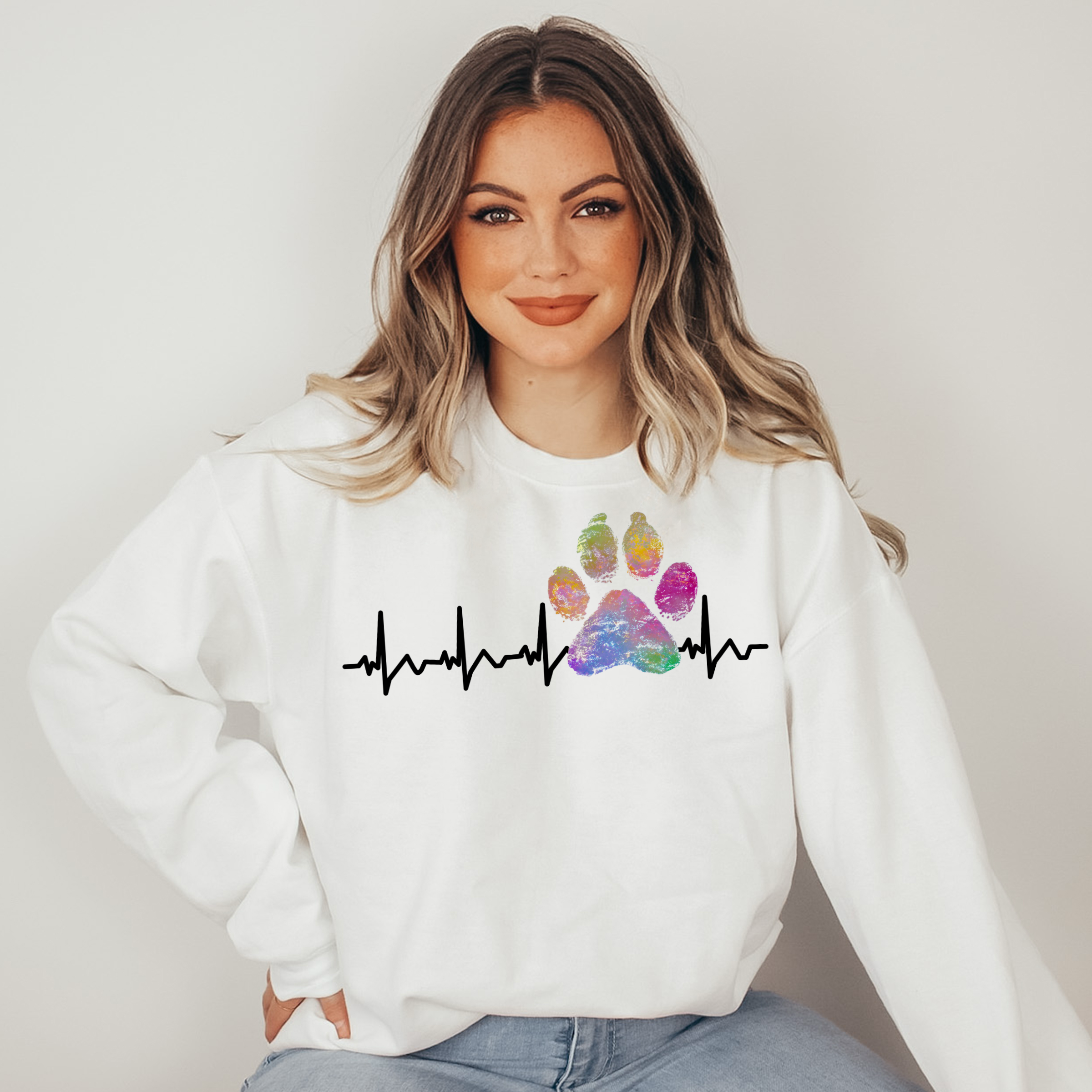 Woman wearing a white sweatshirt with a colorful paw print and heartbeat design on a plain background
