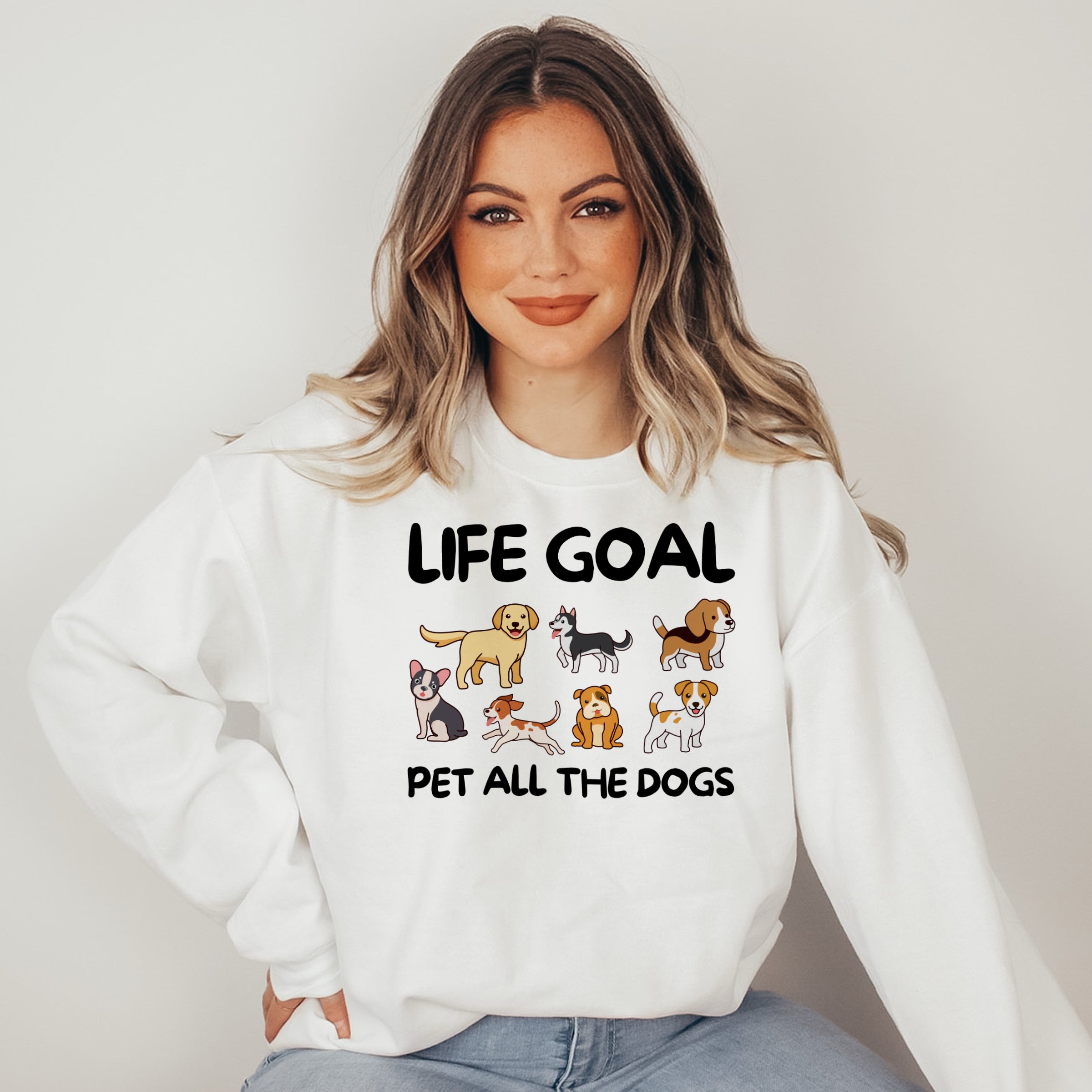 Woman wearing a white sweatshirt with 'Life Goal Pet All The Dogs' text and dog illustrations.