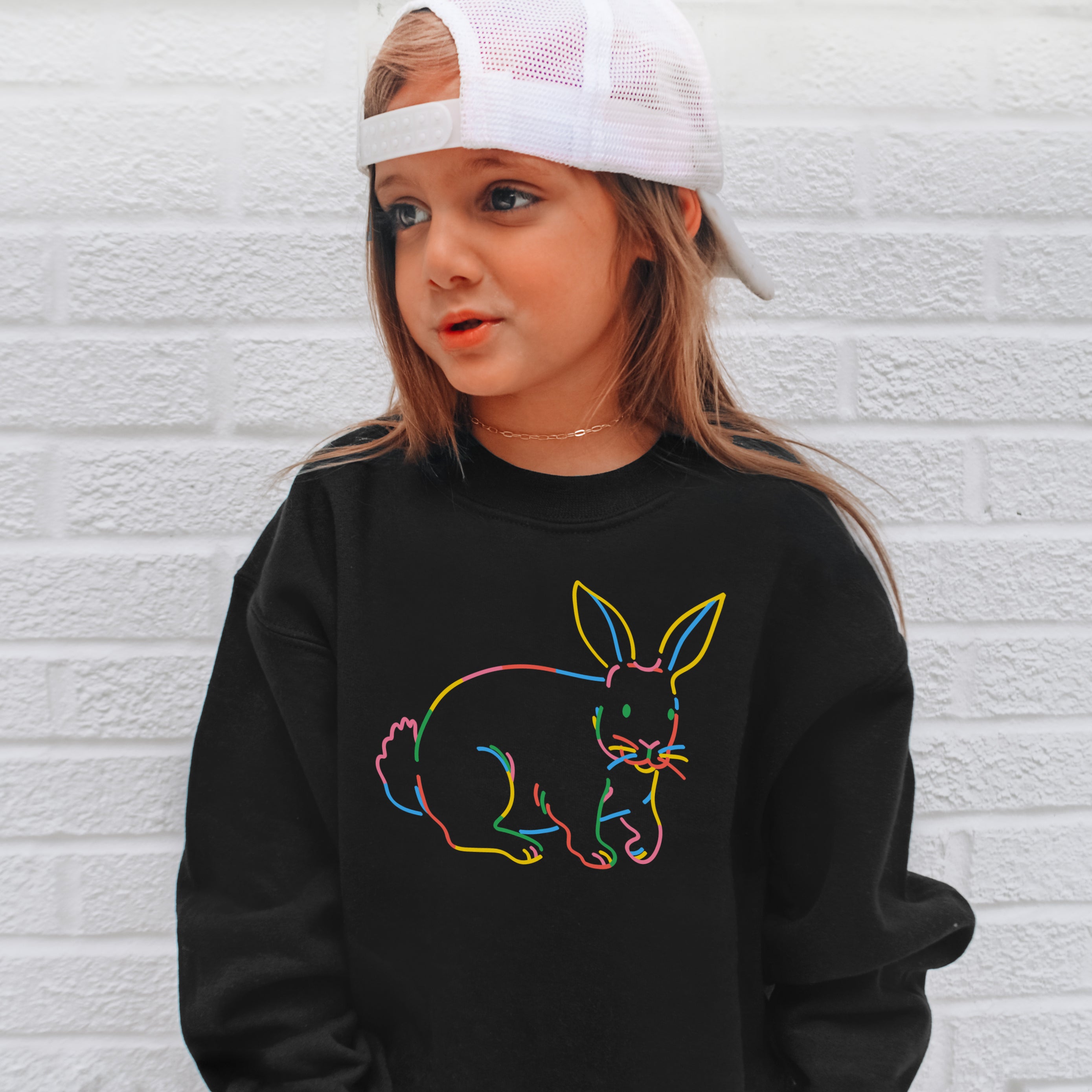 Child wearing a black sweatshirt with a colorful bunny design against a white brick wall.