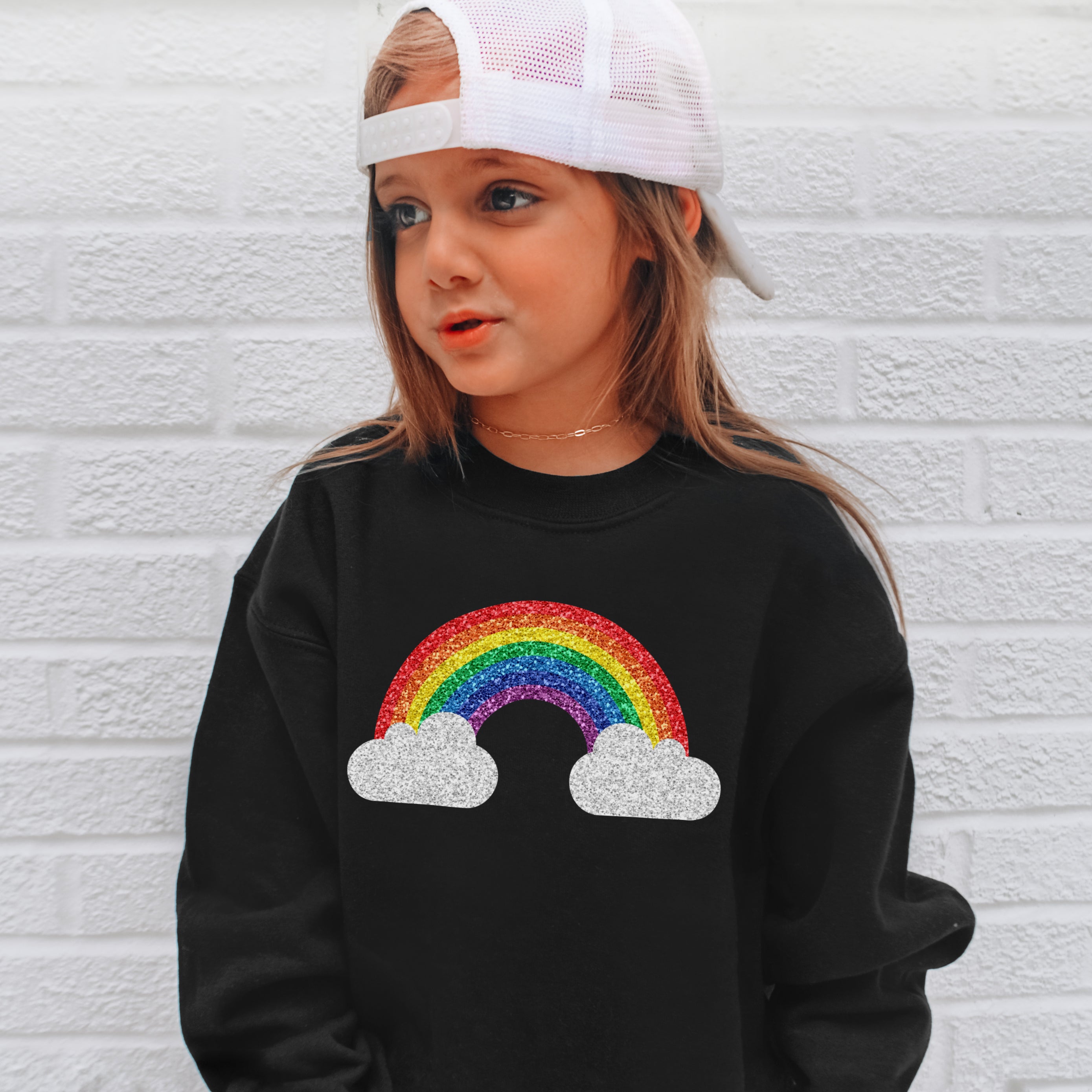 Child wearing a black sweatshirt with a colorful rainbow design against a white brick wall.