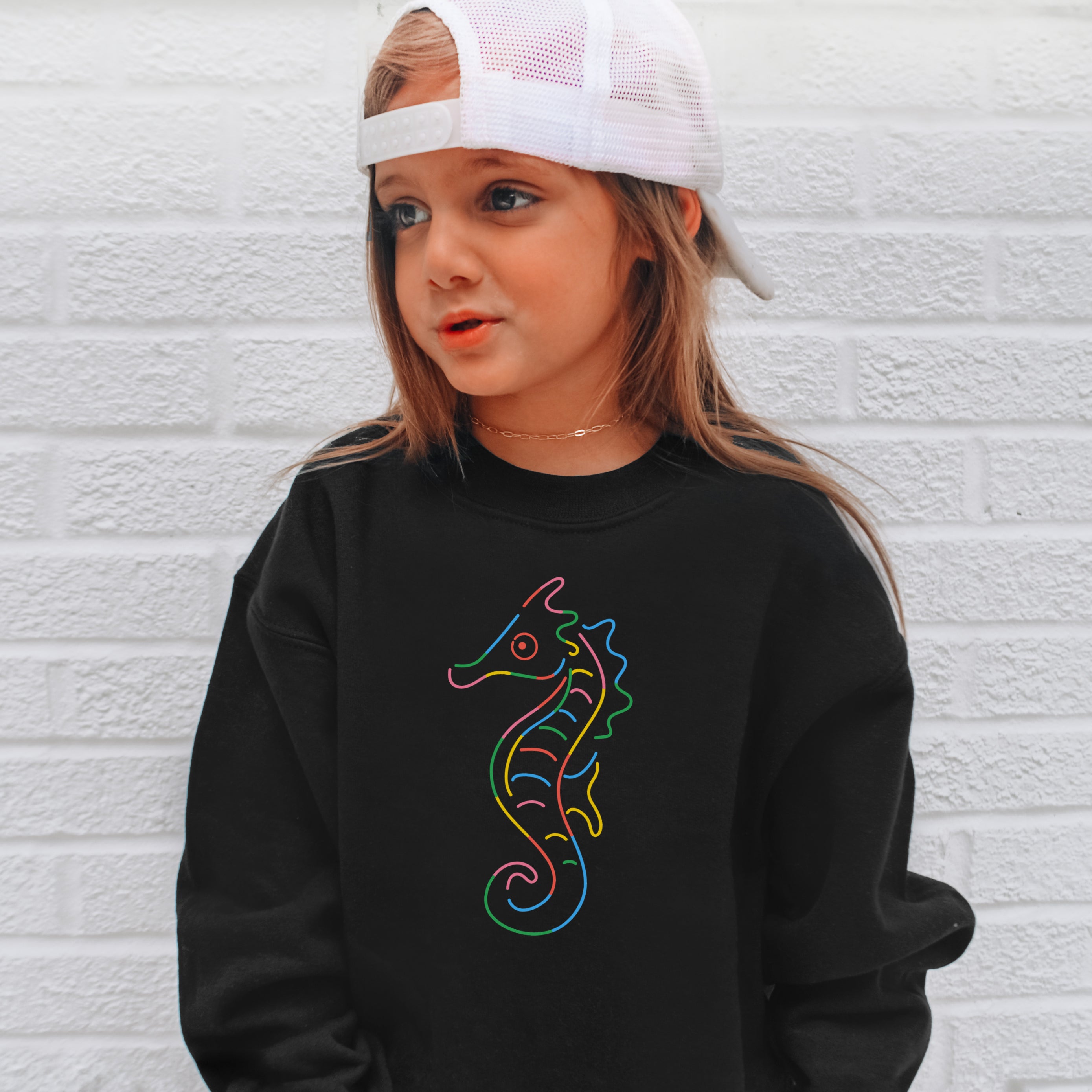 Child wearing a black sweatshirt with a colorful seahorse design against a white brick wall.