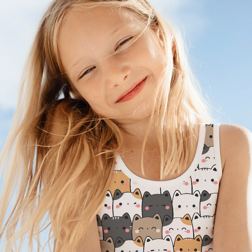 A young girl with long blonde hair smiling outdoors against a bright blue sky, wearing a swimsuit with a colorful cartoon cat pattern.